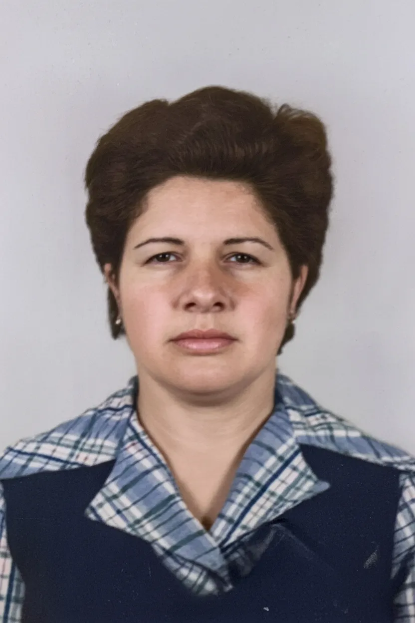 Mulher de meia-idade com cabelo castanho escuro e curto, vestindo uma camisa xadrez azul e branca e um colete azul marinho, olhando diretamente para a frente contra um fundo simples.