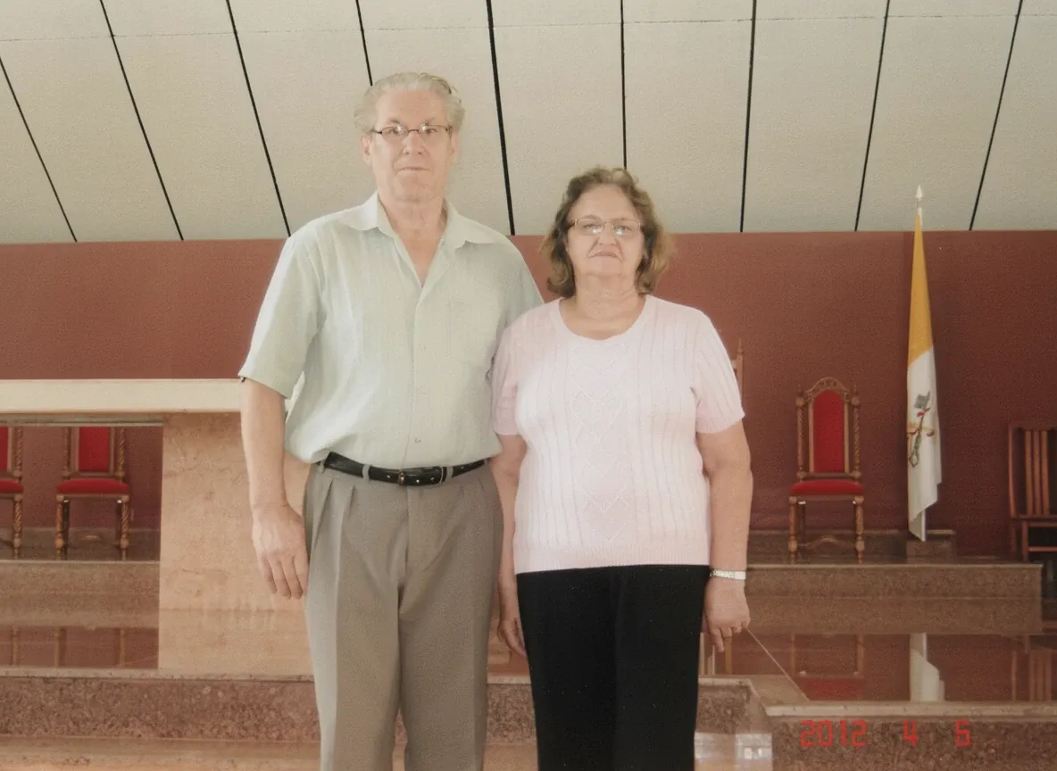 Um homem de meia-idade e uma mulher mais velha ficam lado a lado em um interior espaçoso com tetos altos, paredes vermelhas e uma bandeira amarela e branca.