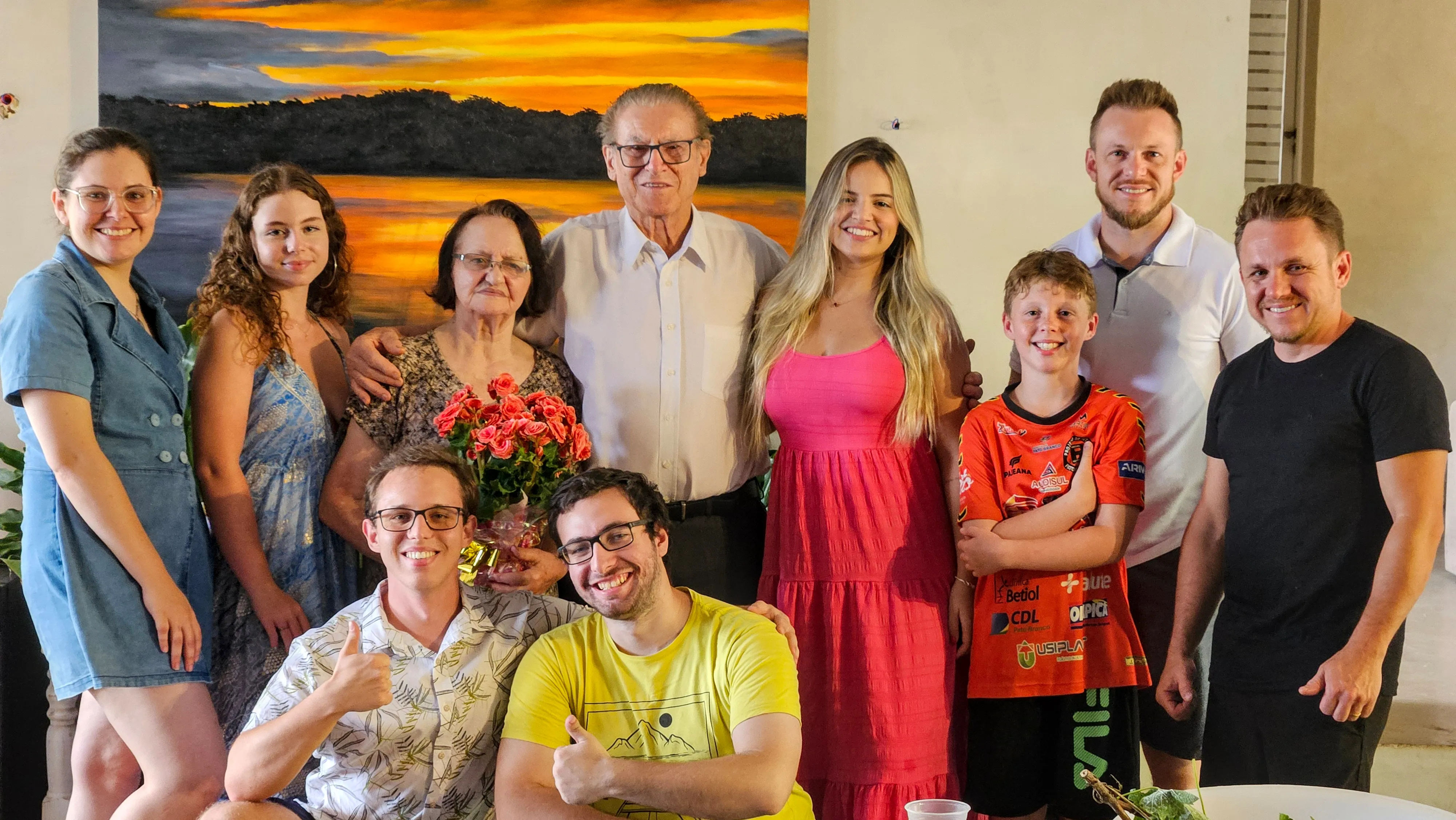 Grupo de dez pessoas, incluindo um homem mais velho e uma mulher mais velha, posando para uma foto em um interior com uma pintura de paisagem pendurada na parede atrás deles.