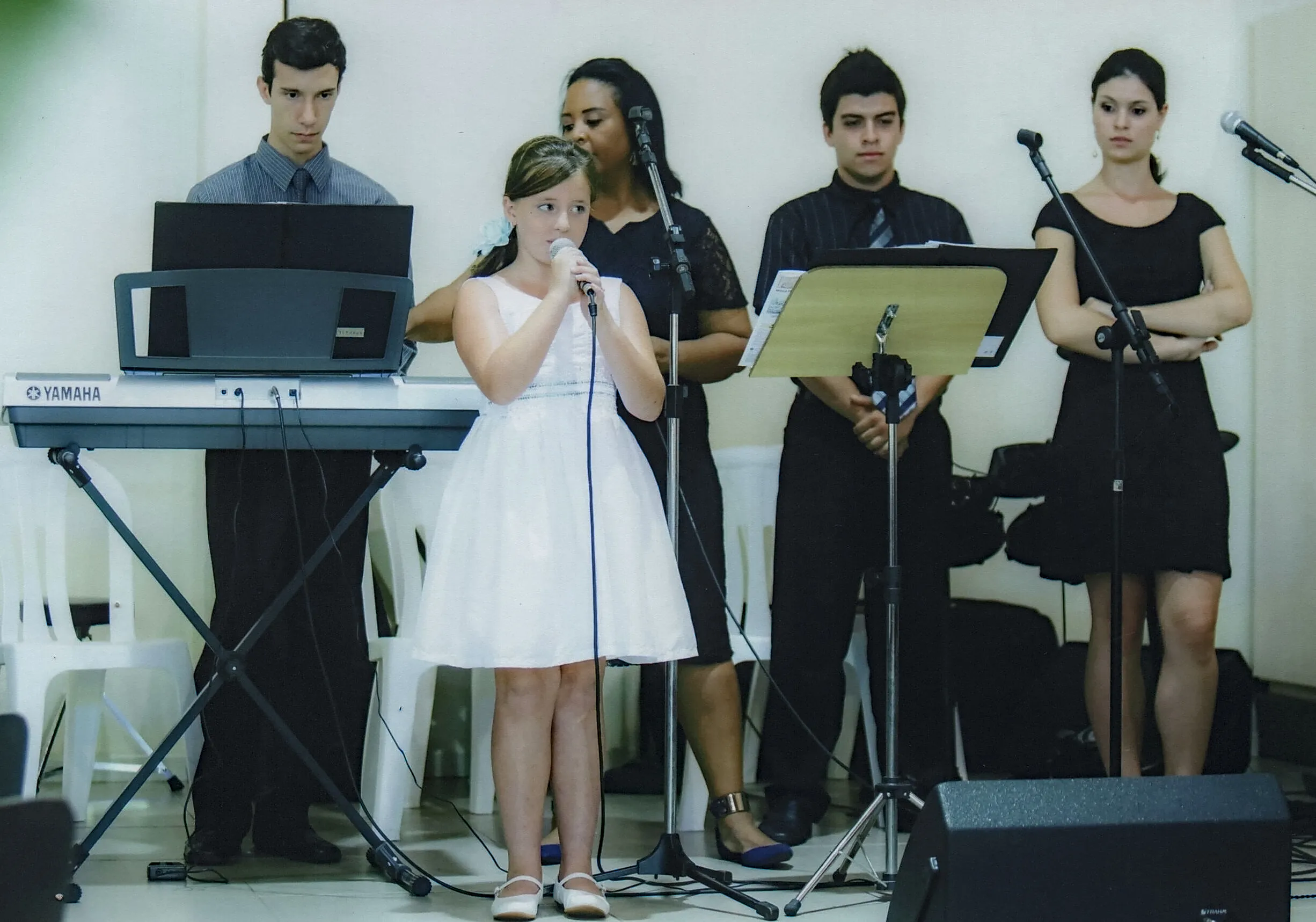 Uma jovem com um vestido branco canta em um microfone no centro, enquanto quatro adultos a observam no palco.