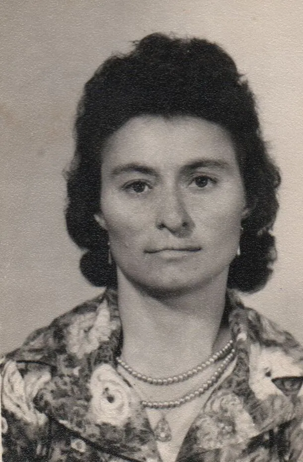 Retrato de uma mulher com cabelo castanho escuro e ondulado, usando um colar duplo de pérolas, brincos dourados e uma blusa floral azul e creme. (Original)