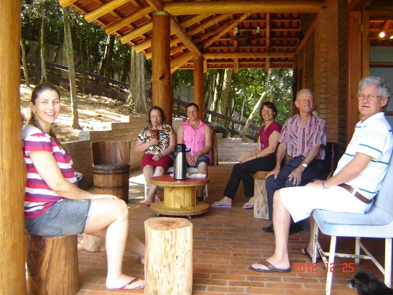 Um grupo de seis adultos e um cão preto estão sentados em um grande pátio de tijolos com telhado de madeira, rodeado por árvores.