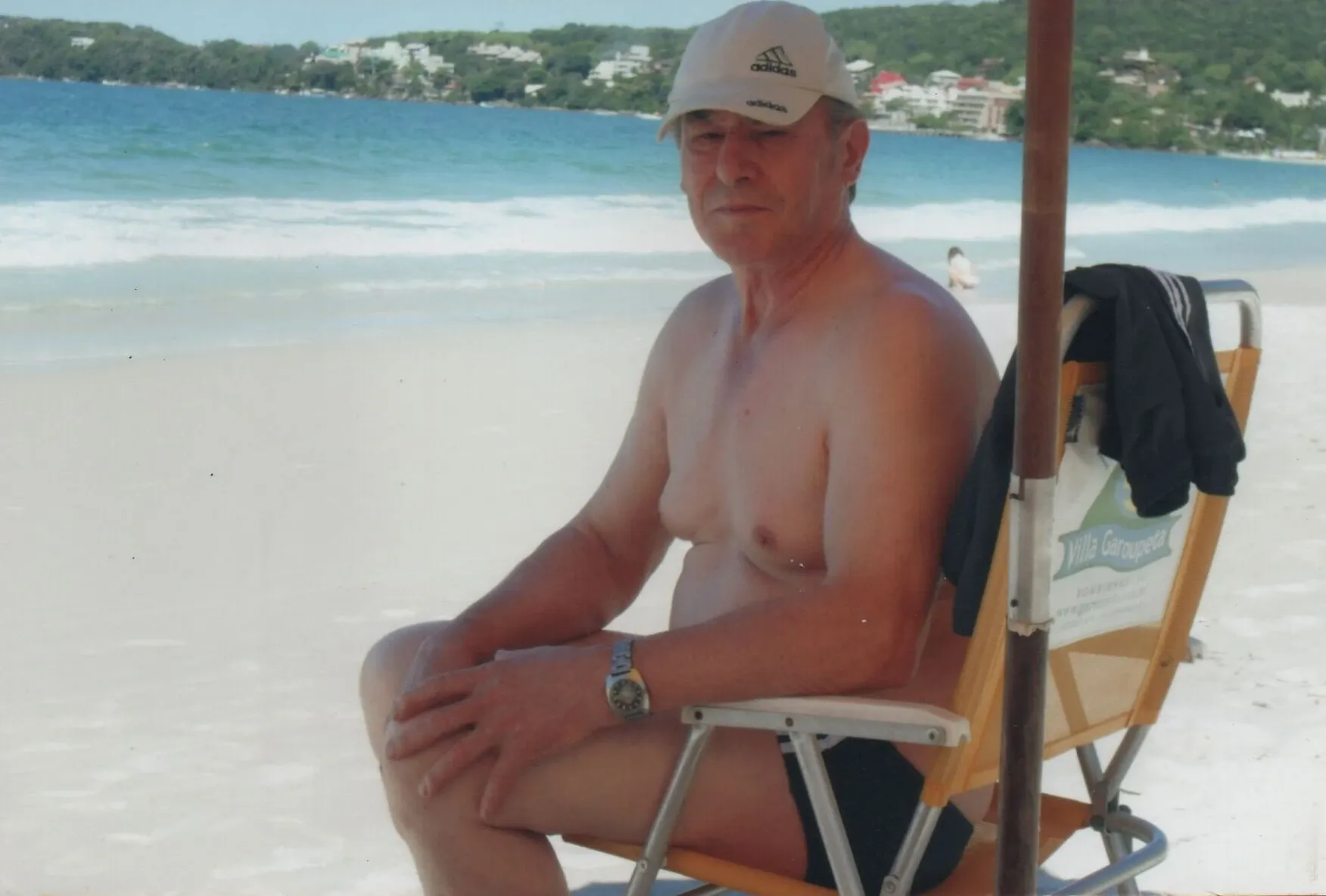 Homem mais velho, de pele clara e de topless, usando boné e sunga preta, sentado em uma cadeira de praia com um guarda-sol na areia branca da praia, olhando para frente.