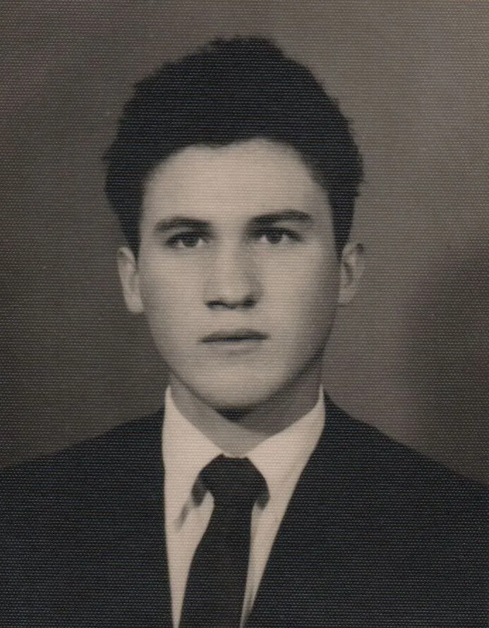 Retrato frontal de um jovem com cabelo castanho escuro e camisa branca de colarinho, gravata azul escura e paletó cinza escuro. (Original)