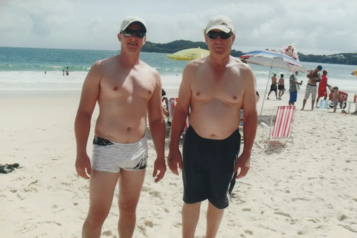 Dois homens caucasianos em trajes de banho e bonés de beisebol, um mais jovem e magro de óculos escuros e calção de banho xadrez, e um mais velho e robusto também de óculos escuros e bermuda preta, estão parados lado a lado na areia da praia, olhando para a frente. O oceano azul e a costa com pessoas e guarda-chuvas estão visíveis ao fundo.