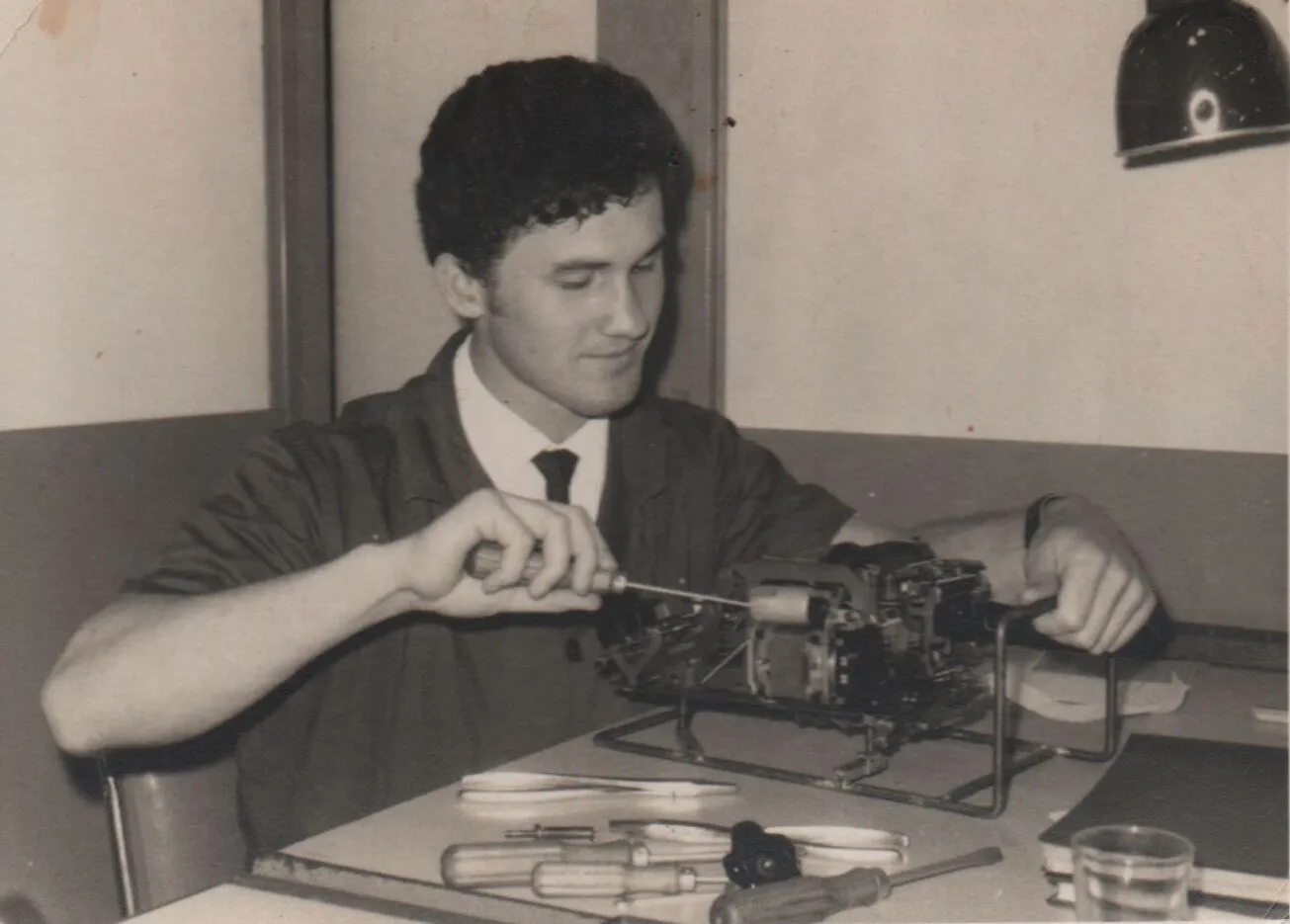Um jovem de cabelo escuro em um uniforme verde-oliva com gravata azul conserta um dispositivo mecânico intrincado em uma mesa de laboratório. (Original)