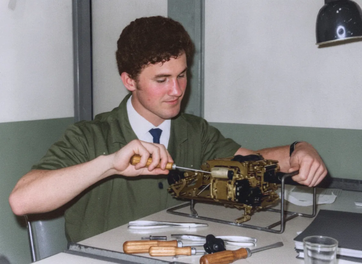 Um jovem de cabelo escuro em um uniforme verde-oliva com gravata azul conserta um dispositivo mecânico intrincado em uma mesa de laboratório.