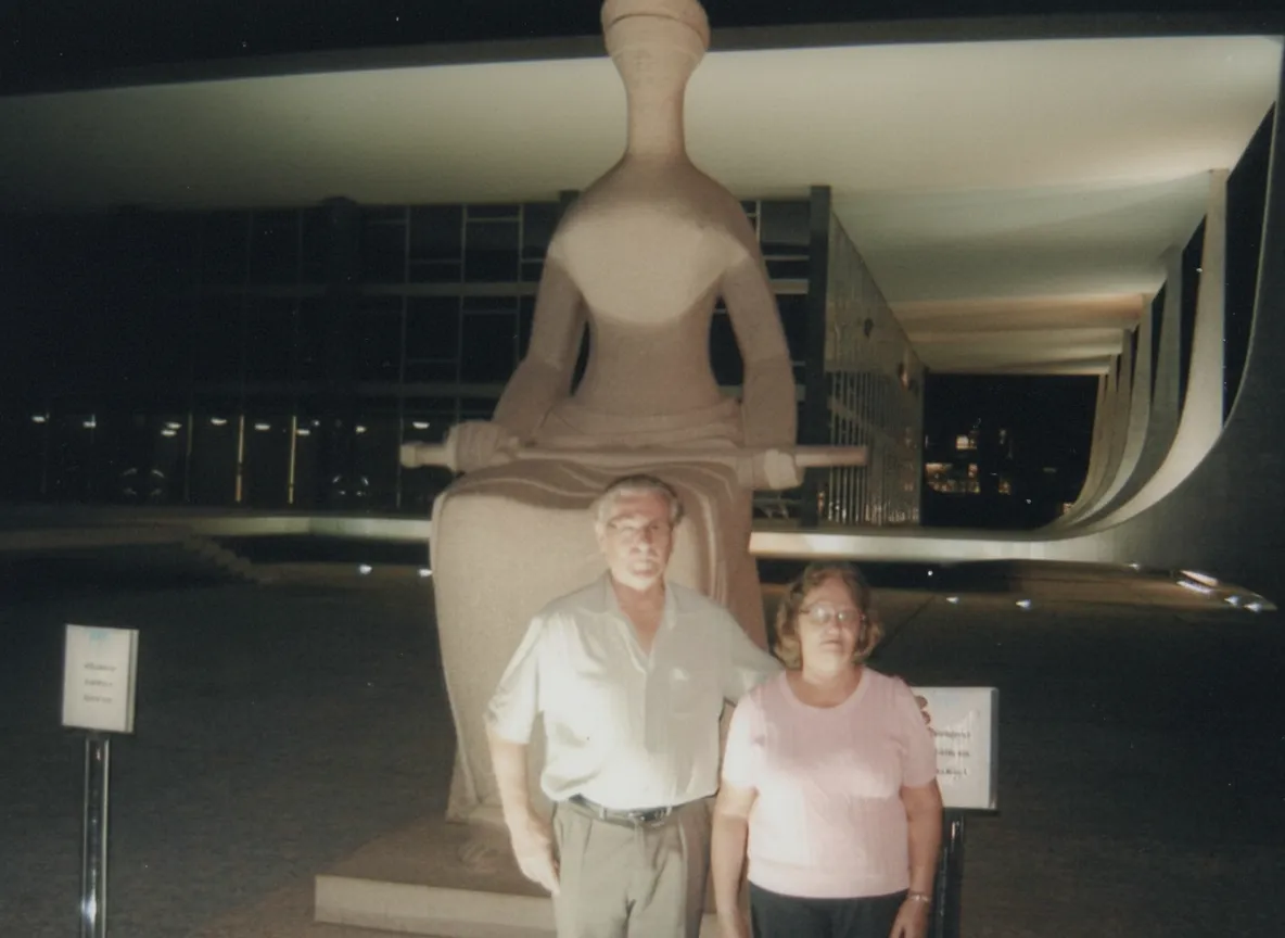 Homem e mulher de meia-idade posam em frente à estátua de A Justiça no Supremo Tribunal Federal de Brasília, à noite.