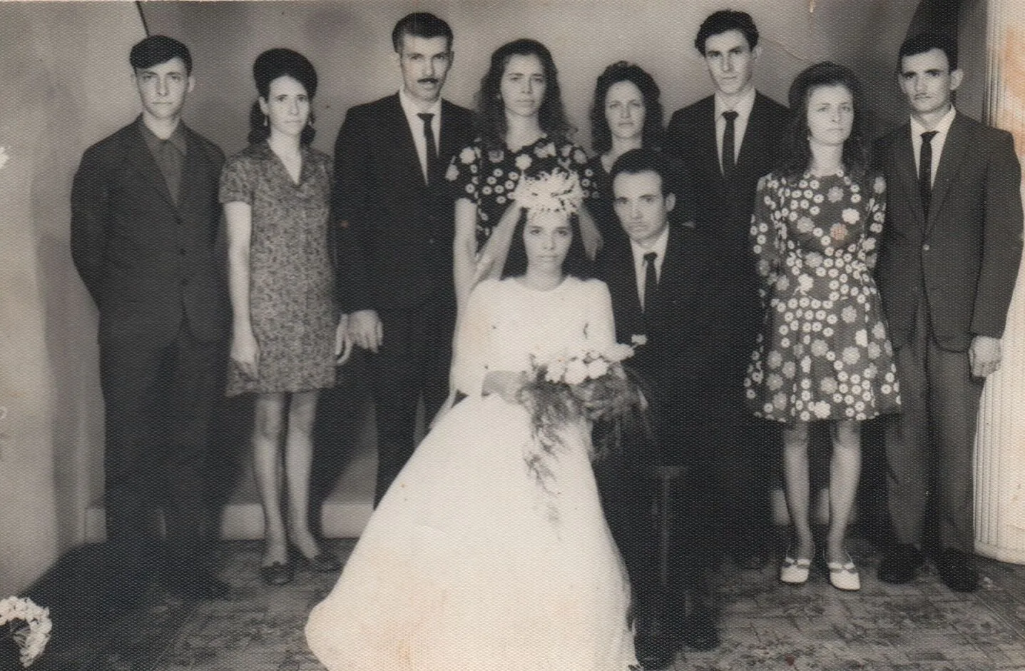 Foto de casamento de 1960 com dez pessoas, com a noiva sentada na frente ao lado do noivo, e os convidados, homens de terno e mulheres de vestido, emoldurando o casal. (Original)