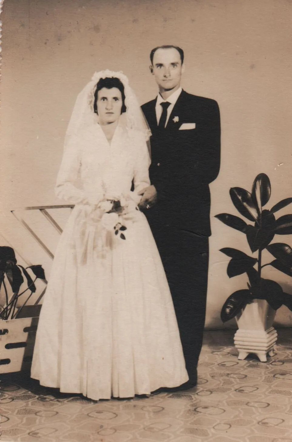 Casal de noivos posa para foto: ela, de vestido branco e véu, segura buquê; ele, de terno azul escuro, está ao lado dela. (Original)