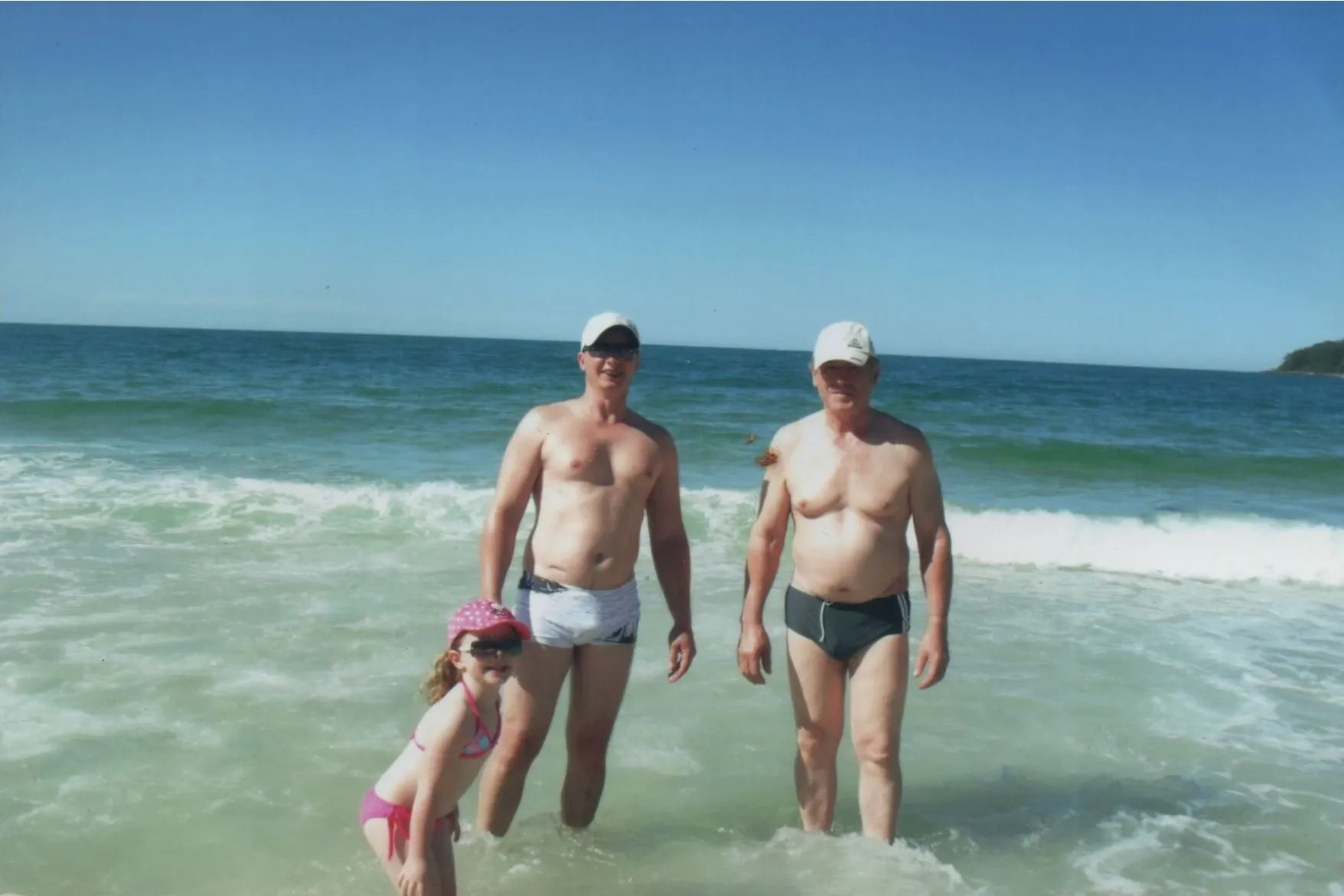 Dois homens, um jovem e um mais velho, e uma criança, estão na água do oceano, sorrindo para a câmera.