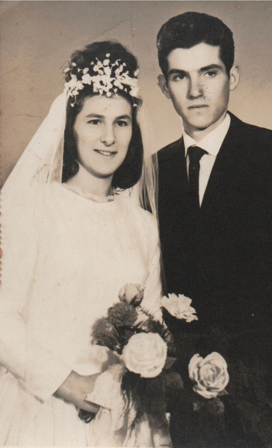 Retrato de um casal em trajes de casamento; a noiva sorri, segurando um buquê de rosas, ao lado do noivo. (Original)