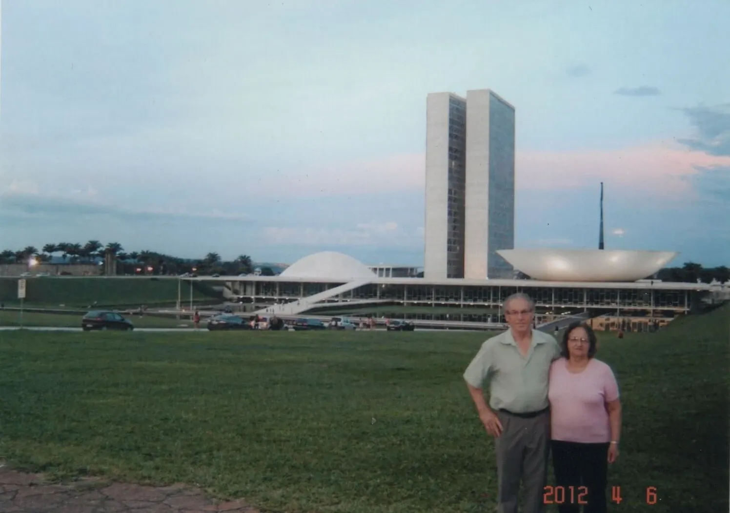 Um casal de idosos em pé em frente ao Congresso Nacional do Brasil, com um céu nublado ao entardecer.