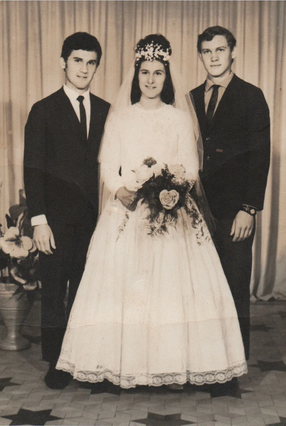 Noiva sorrindo com véu e buquê, ladeada por dois homens de terno, todos olhando para frente em uma foto de casamento vintage. (Original)