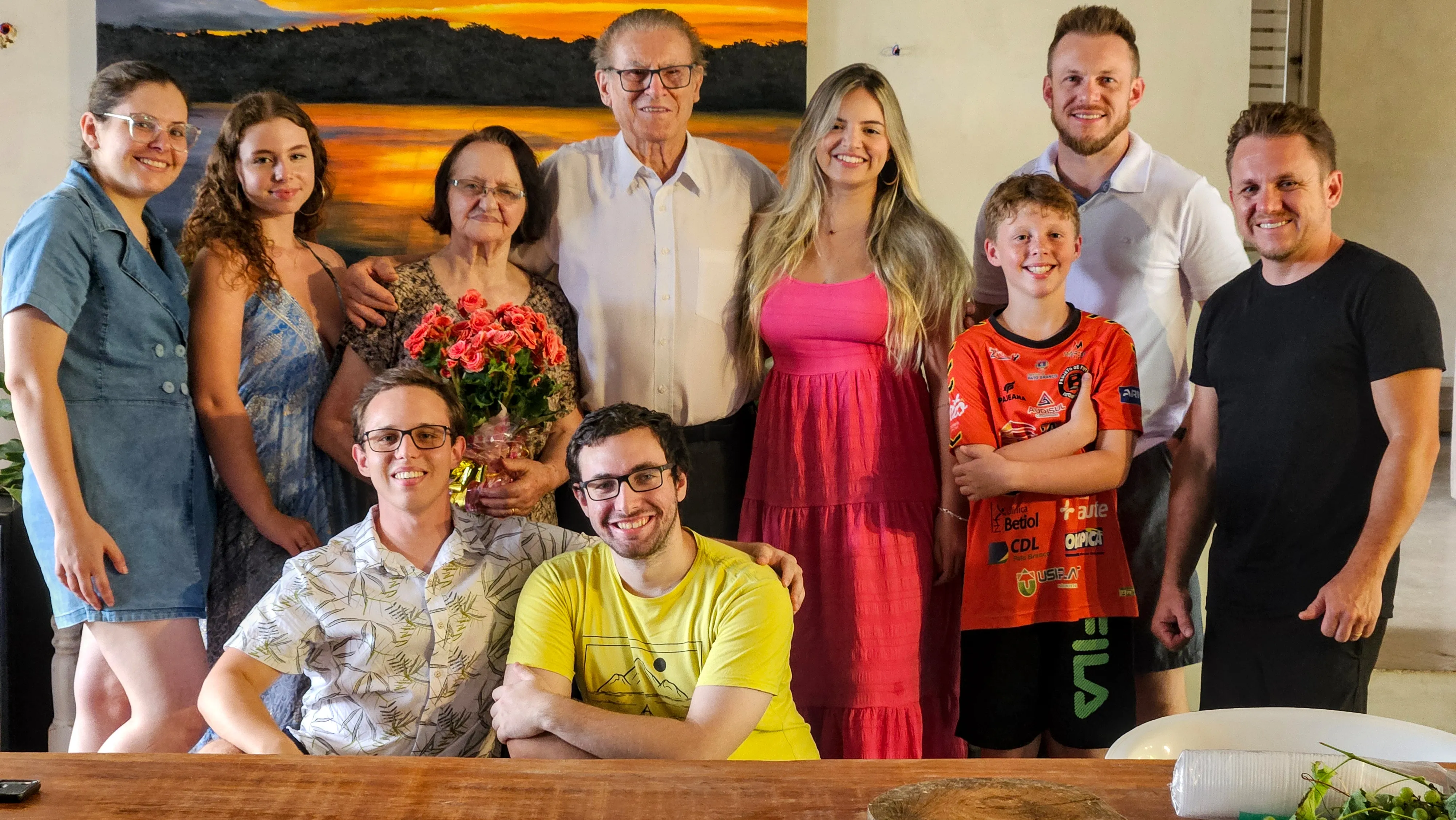 Uma família de dez pessoas de diferentes idades posa para uma foto, em pé e sentadas, sorrindo em um quarto com uma pintura de paisagem pendurada na parede atrás delas.