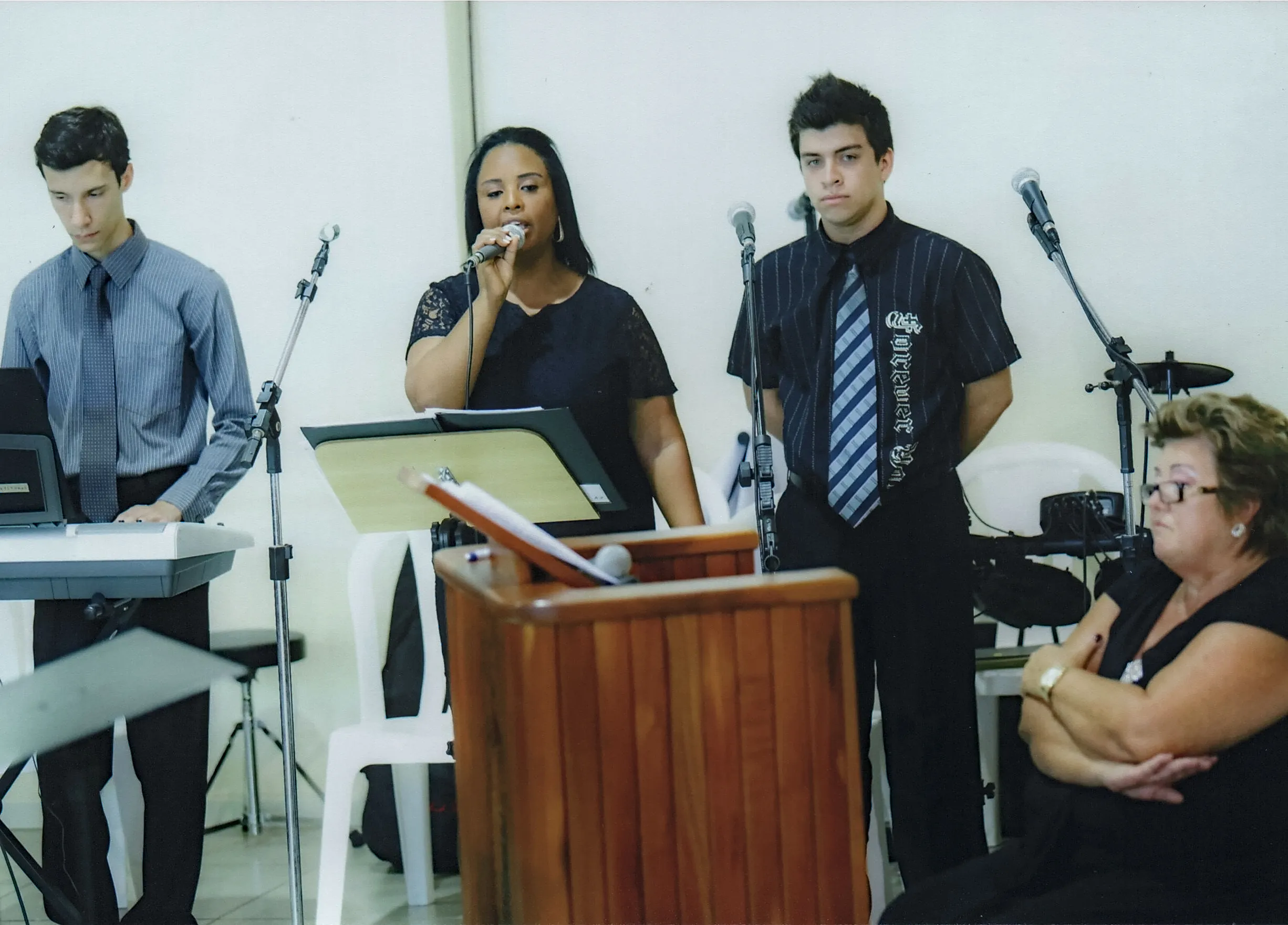 Uma mulher em pé no centro cantando em um microfone no pódio, ladeada por dois homens e uma mulher sentada à direita.