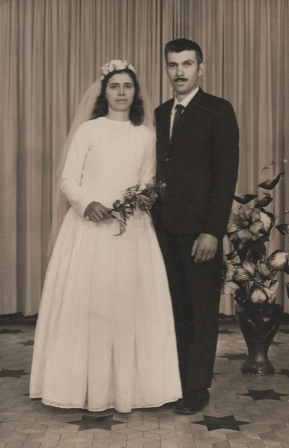 Casamento: Noiva séria em um vestido branco e coroa de flores, segurando um buquê, ao lado de um noivo sério de terno escuro. (Original)