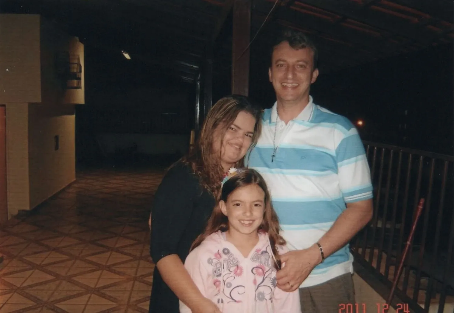 Uma família de três pessoas, um homem, uma mulher e uma menina, estão sorrindo para a câmera enquanto estão lado a lado, o homem abraçando a menina e a mulher.