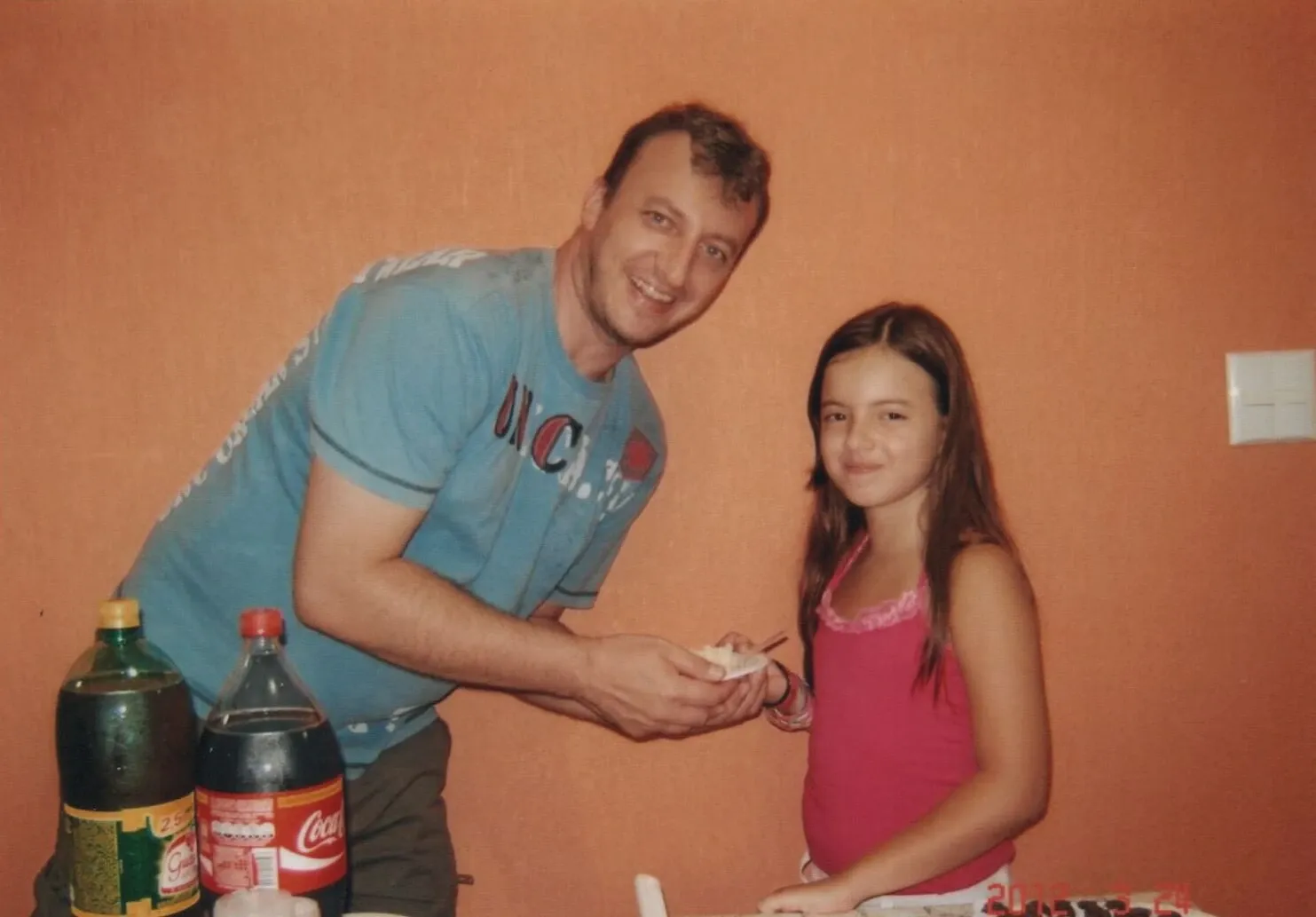 Homem em uma camiseta azul com estampa branca, sorrindo e oferecendo um pedaço de bolo a uma menina em uma camiseta regata rosa, em frente a uma parede laranja.