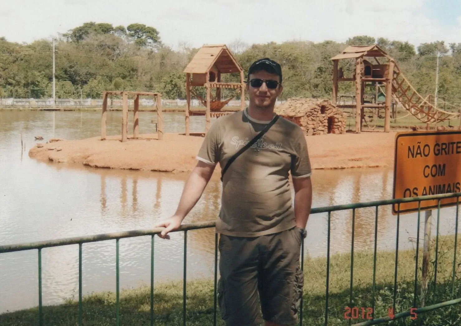 Um homem em uma camiseta marrom-esverdeada, shorts e óculos escuros está em pé na frente de uma cerca verde, apoiando-se nela com a mão esquerda, com um lago e cabanas de madeira à sua direita, sob um céu claro.