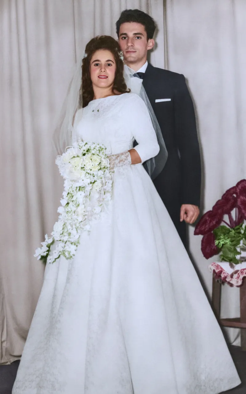 Uma noiva sorridente em um vestido branco e véu segura um buquê de flores enquanto está ao lado de um noivo sério de smoking.