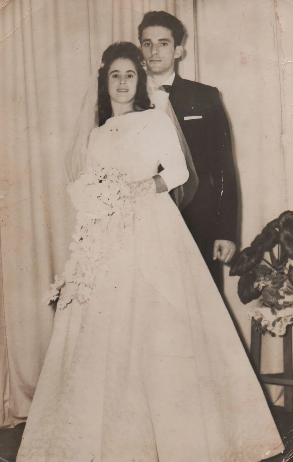 Uma noiva sorridente em um vestido branco e véu segura um buquê de flores enquanto está ao lado de um noivo sério de smoking. (Original)