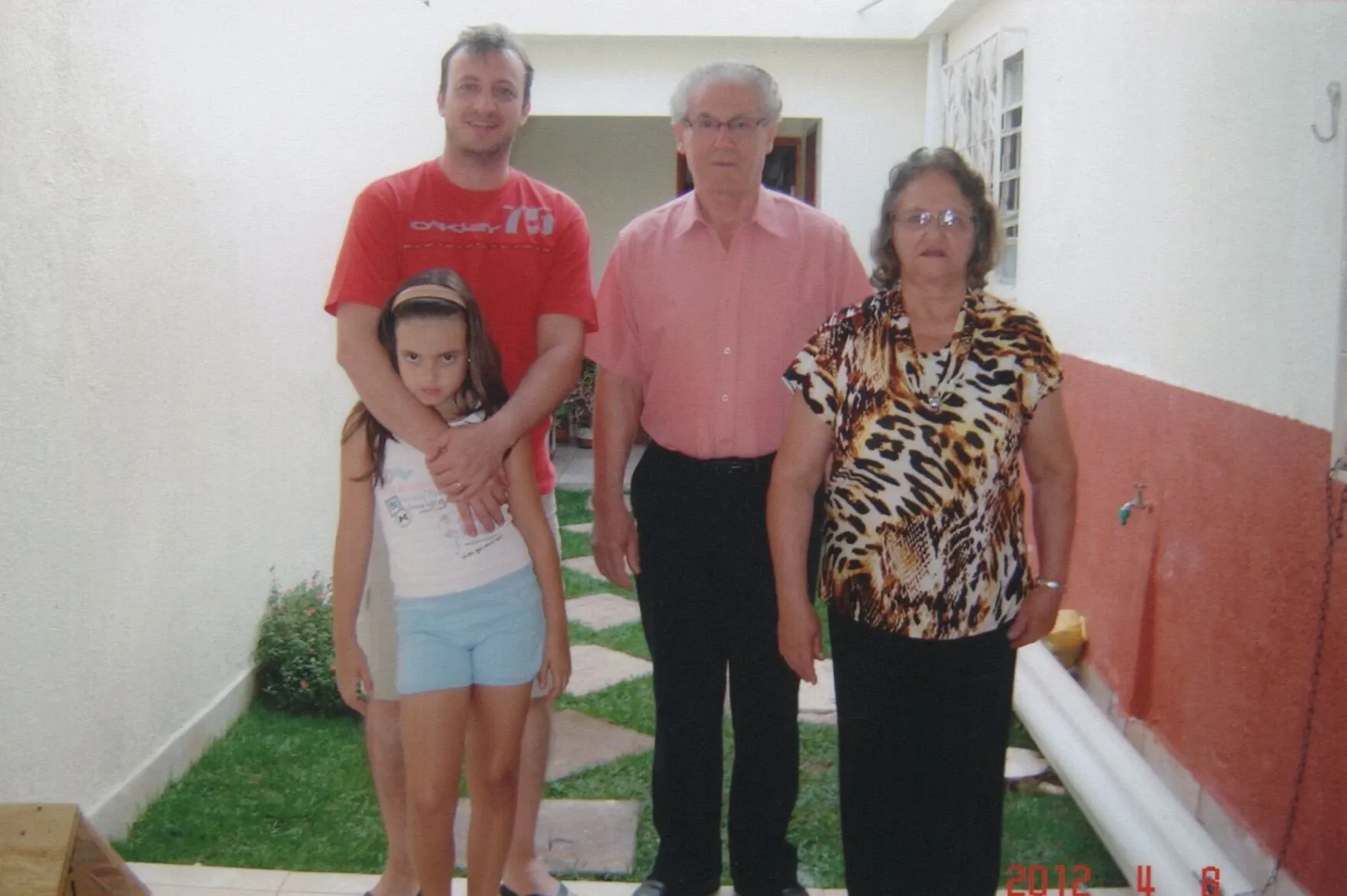 Quatro indivíduos de três gerações: um homem, um homem idoso, uma mulher idosa e uma menina, em pé em um corredor estreito com paredes brancas e grama verde no chão.