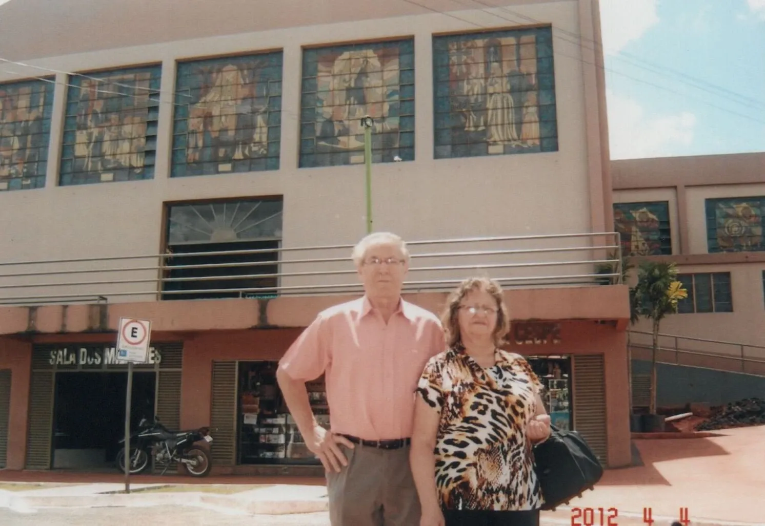 Um homem de camisa rosa e calças cinzas, e uma mulher de blusa estampada de leopardo e calça preta, posam para uma foto em frente a um prédio com janelas coloridas e uma placa "SALA DOS MUSEUS".