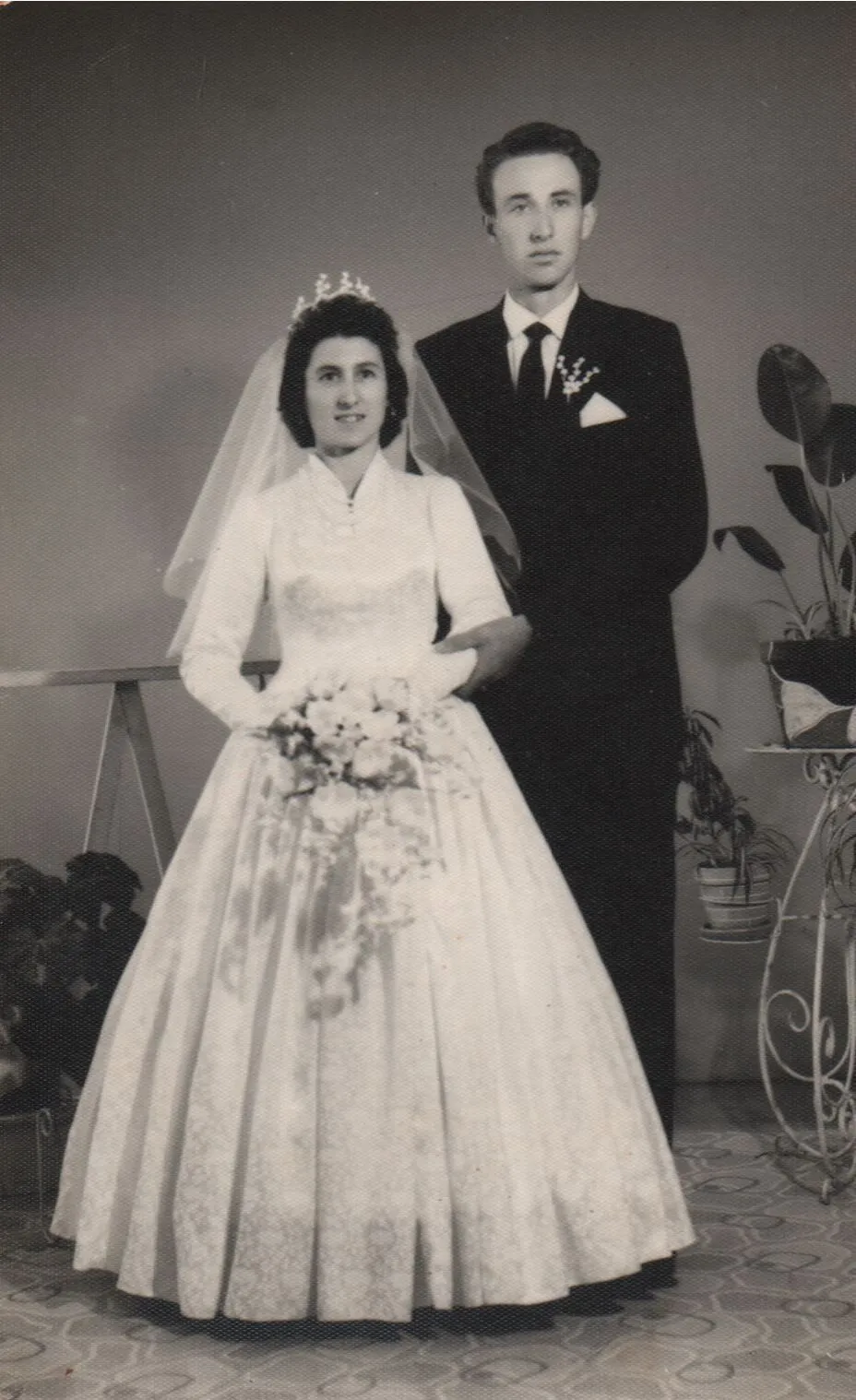 Retrato de casamento de um casal: a noiva está em primeiro plano, usando um vestido de cetim branco com mangas compridas e um véu, segurando um buquê de flores. O noivo está atrás dela, usando um terno escuro e gravata. (Original)