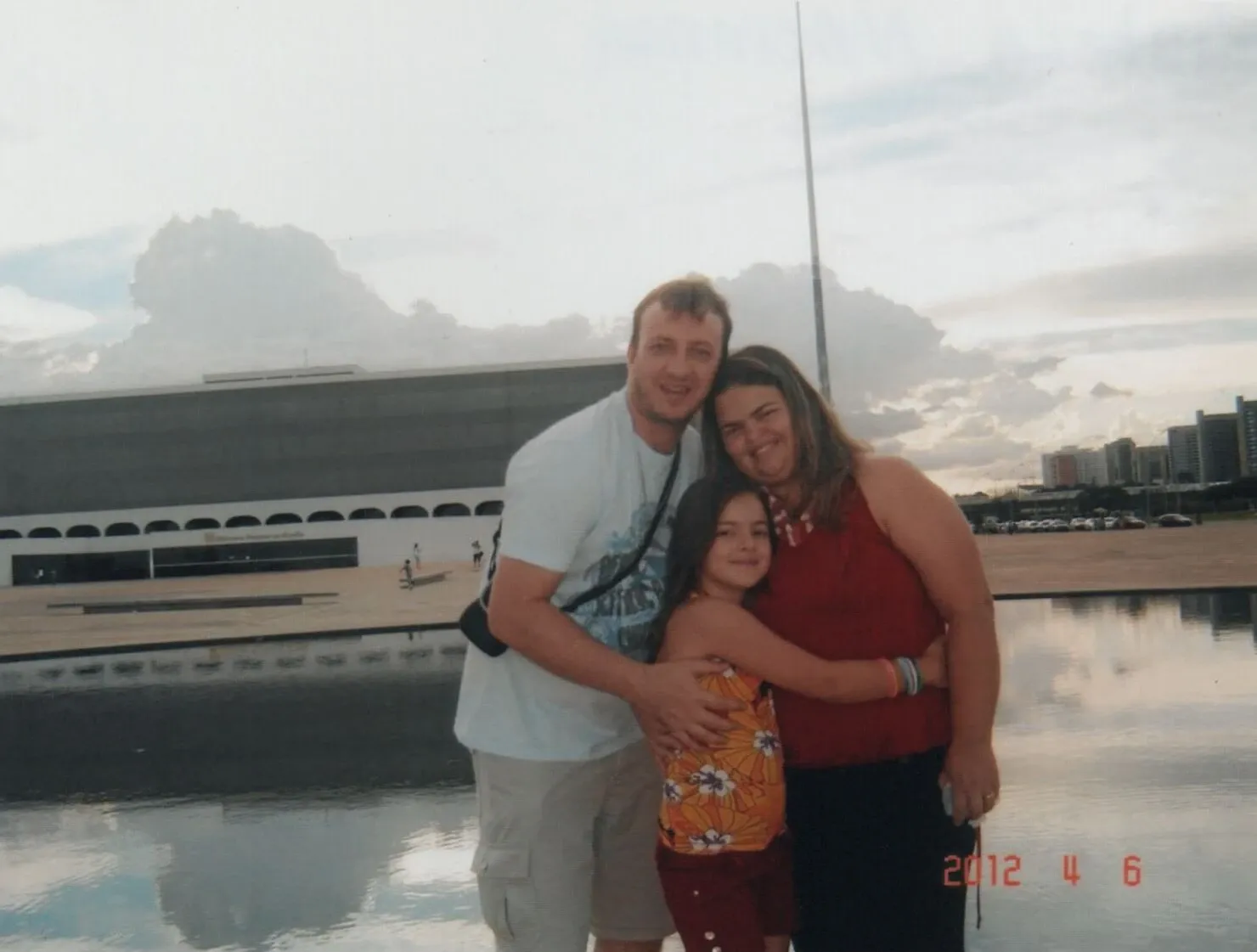 Uma família de três pessoas, incluindo um homem, uma mulher e uma criança, posa para uma foto do lado de fora de um edifício grande e moderno com um corpo de água refletindo nuvens brancas, sob um céu claro e ensolarado.