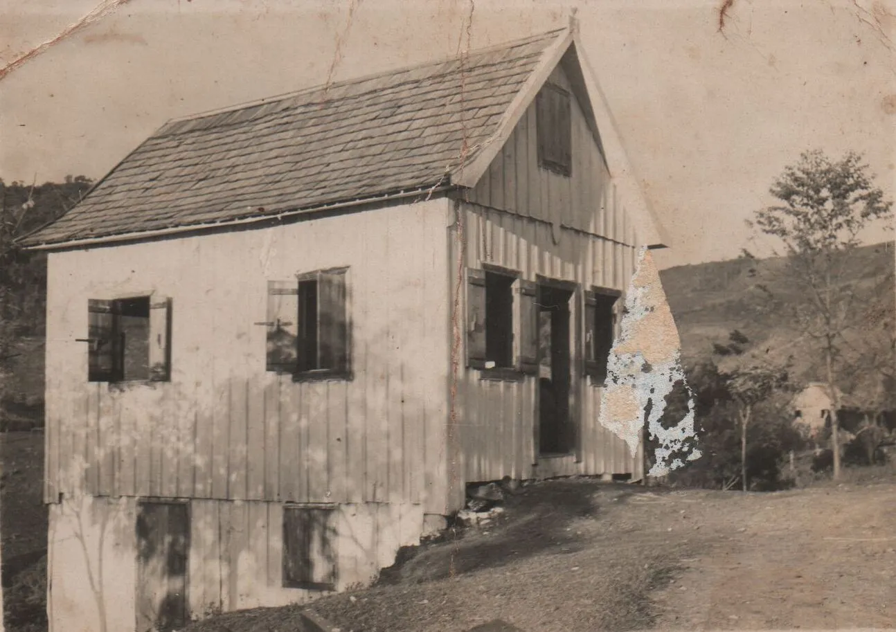 Uma casa de madeira branca e envelhecida com telhado de terracota fica no topo de uma colina de terra batida, com janelas e portas com venezianas azuis abertas, e uma paisagem montanhosa distante sob um céu azul claro. (Original)