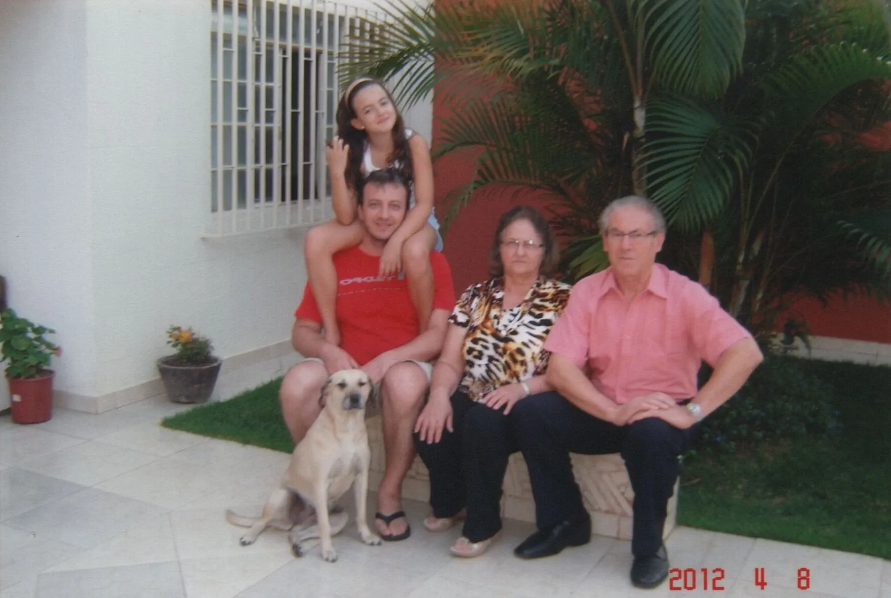 Um homem com uma menina nos ombros, uma mulher e um homem mais velho estão sentados em um banco ao ar livre com um cachorro ao lado deles, em frente a uma casa.