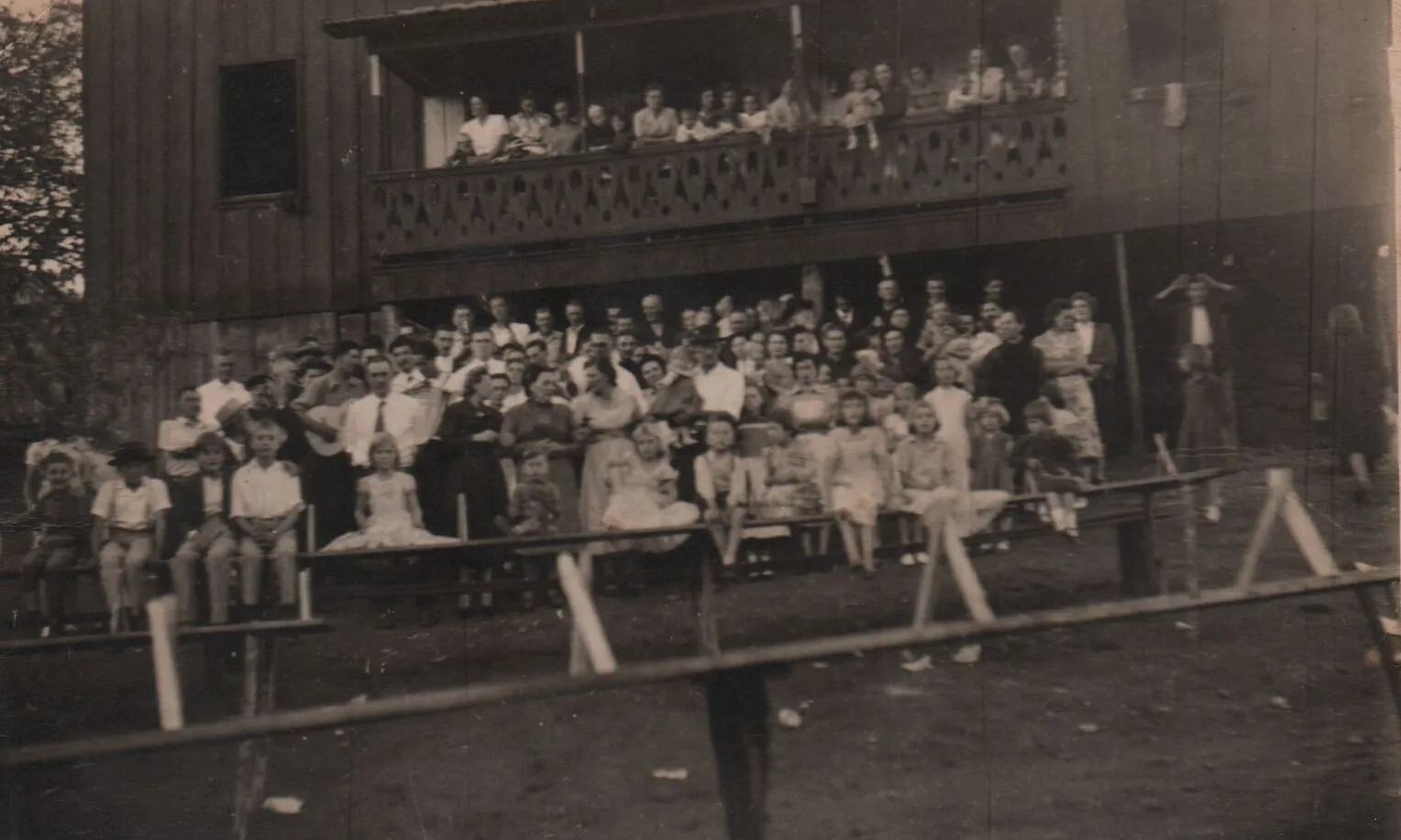 Uma fotografia sépia de um grande grupo de pessoas, incluindo muitos adultos e crianças, reunidas na varanda e em frente a um prédio de madeira de dois andares com uma varanda verde escura. (Original)