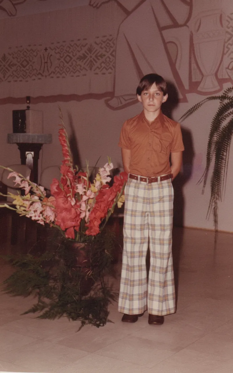 Um jovem com cabelo castanho e camisa polo cor de ferrugem está em pé ao lado de um grande vaso de flores vermelhas e brancas, usando calças xadrez brancas e marrons e sapatos marrons. (Original)