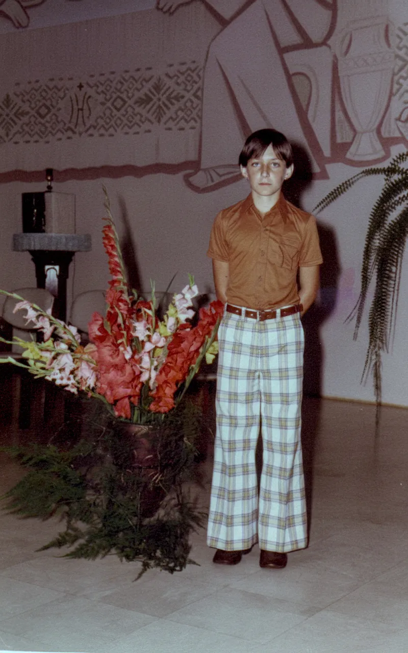 Um jovem com cabelo castanho e camisa polo cor de ferrugem está em pé ao lado de um grande vaso de flores vermelhas e brancas, usando calças xadrez brancas e marrons e sapatos marrons.