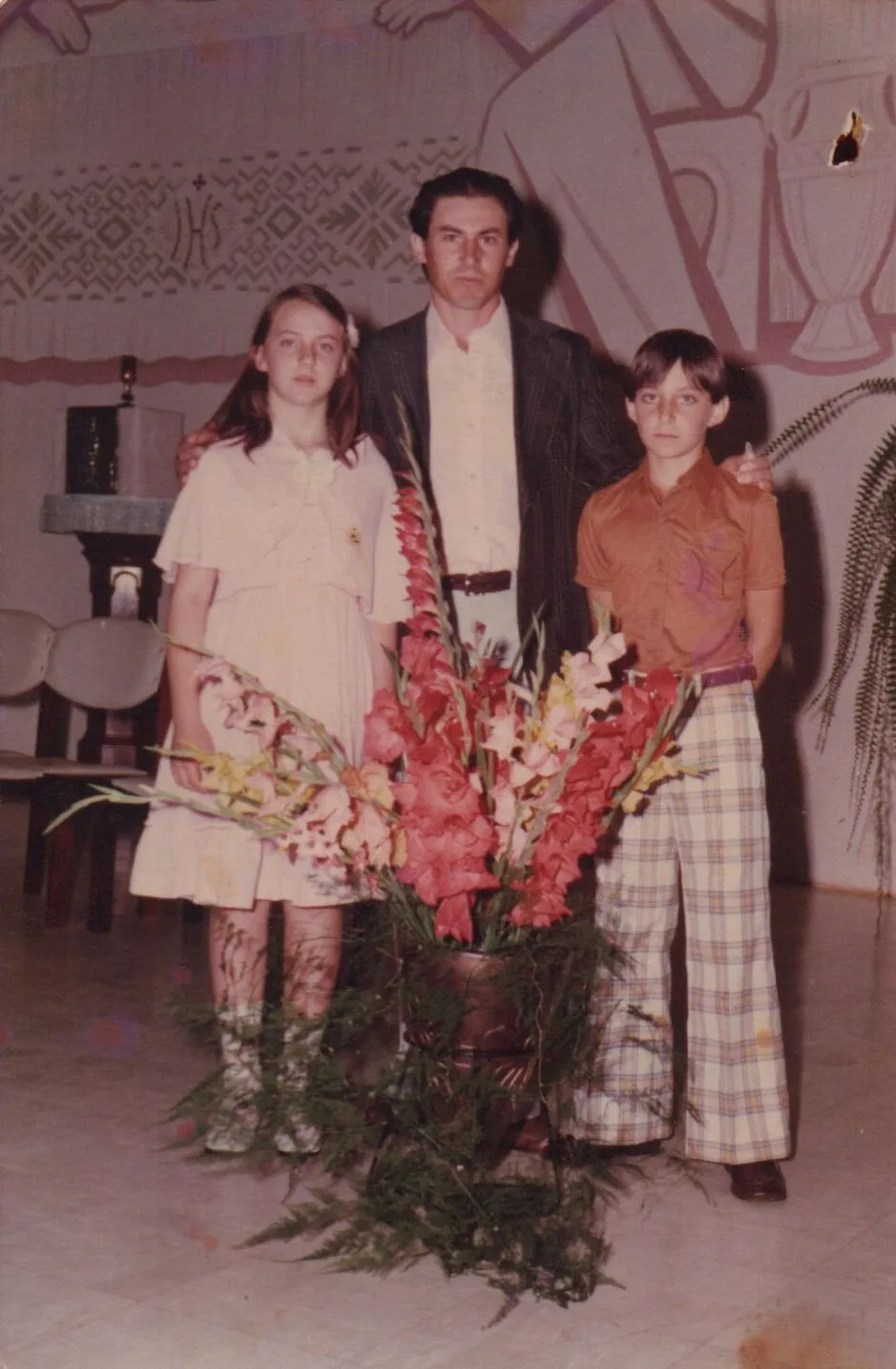 Um homem, uma menina e um menino em pé atrás de um vaso de flores em um palco com um mural abstrato ao fundo. (Original)