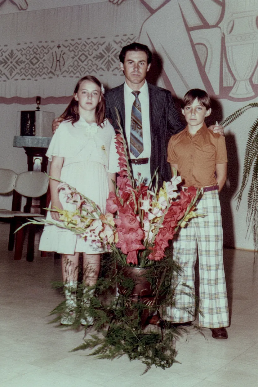 Um homem, uma menina e um menino em pé atrás de um vaso de flores em um palco com um mural abstrato ao fundo.