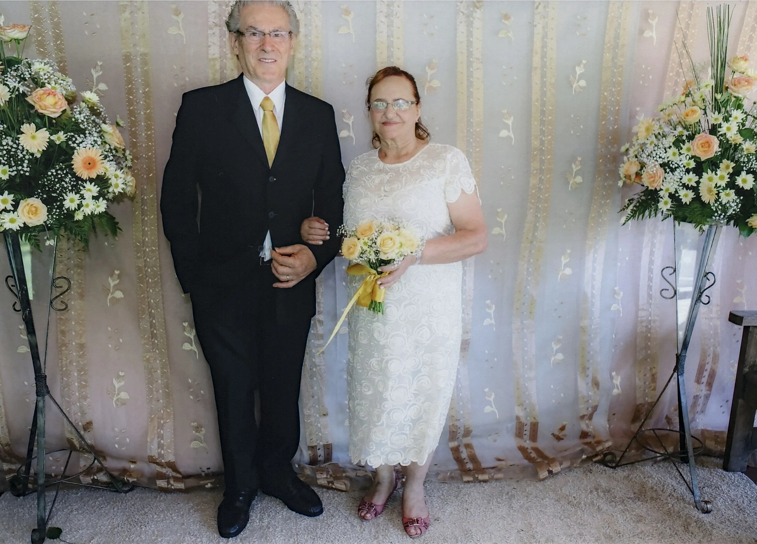 Um casal mais velho está vestido para uma cerimônia de casamento. A mulher usa um vestido branco de renda e segura um buquê de rosas amarelas e flores brancas, enquanto o homem está de terno preto e gravata dourada, com o braço em volta dela.