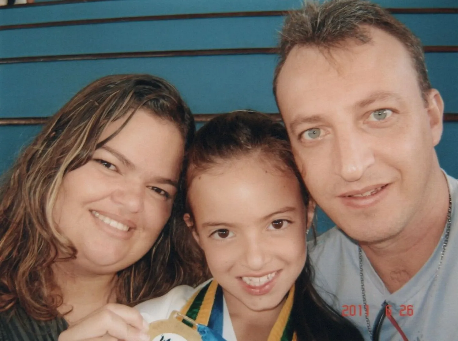 Uma família sorridente posa para uma selfie, com uma garota no centro exibindo uma medalha, possivelmente celebrando uma conquista.