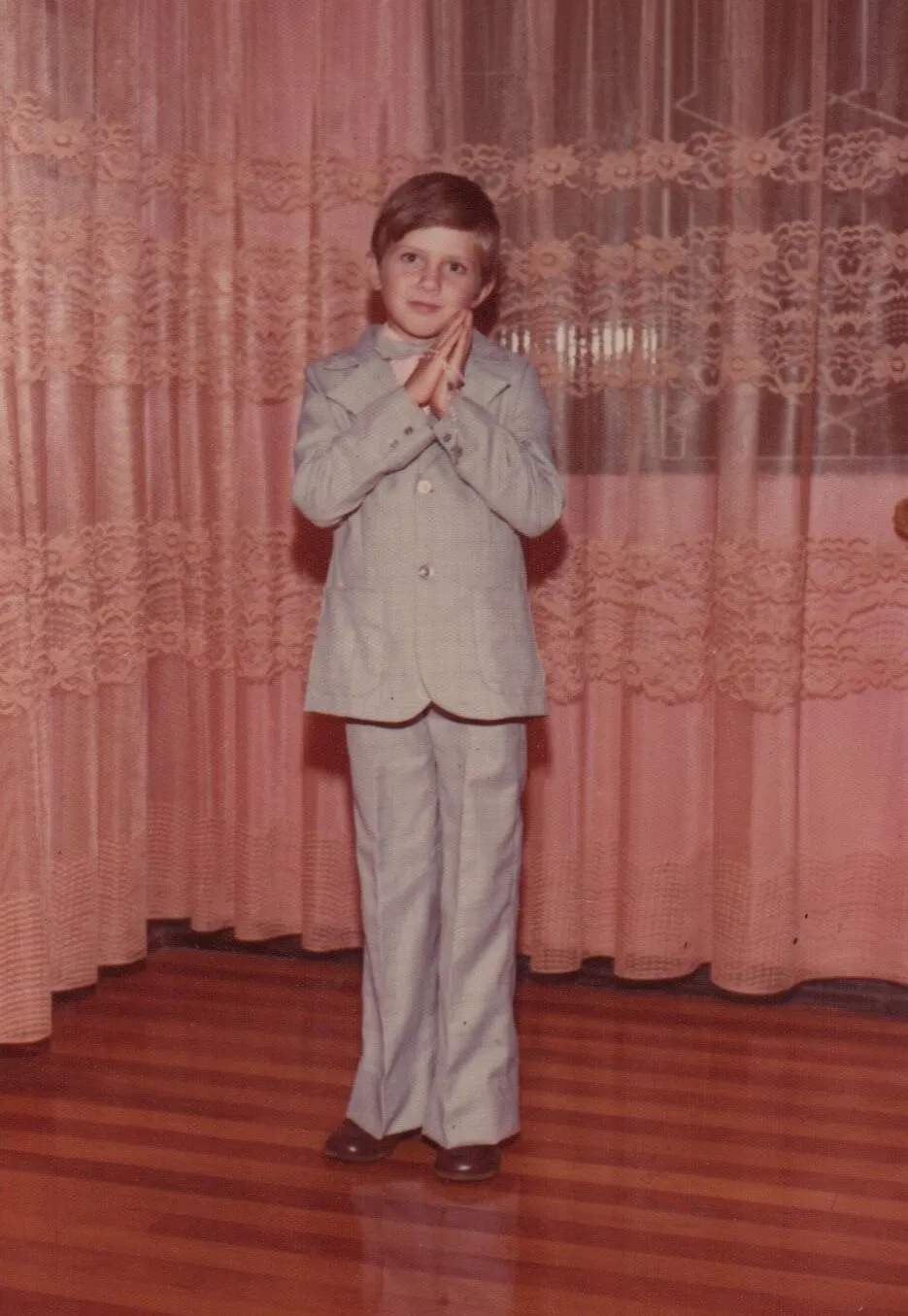 Um menino jovem com cabelo castanho e um terno cinza-claro em um ambiente interno, com as mãos cruzadas na frente do peito, em pé diante de cortinas de renda de cor pêssego e um piso de madeira. (Original)