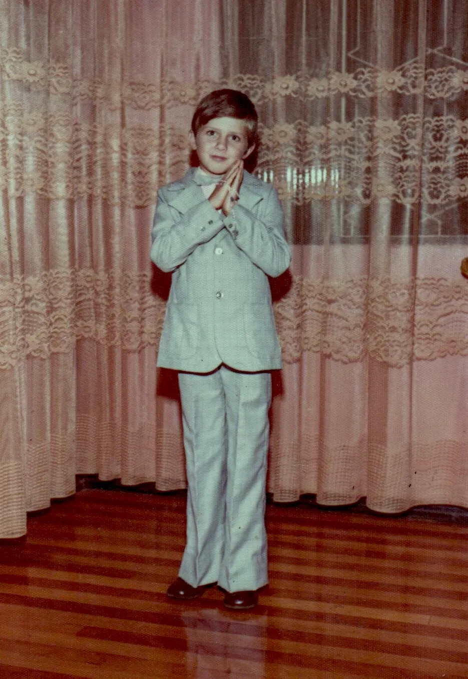 Um menino jovem com cabelo castanho e um terno cinza-claro em um ambiente interno, com as mãos cruzadas na frente do peito, em pé diante de cortinas de renda de cor pêssego e um piso de madeira.