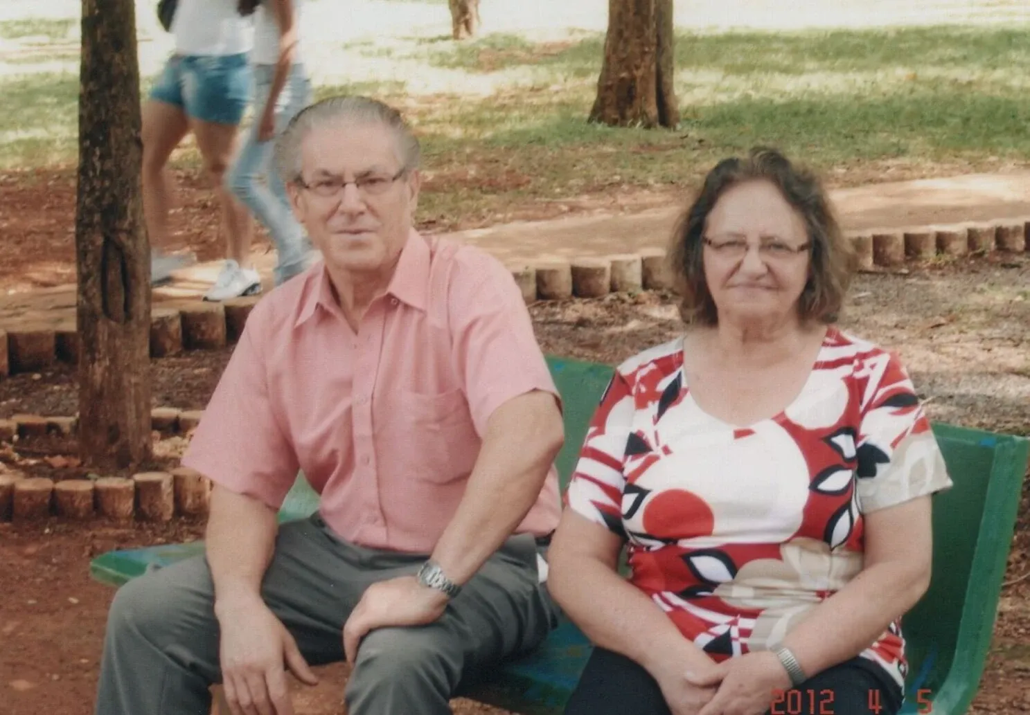 Um casal de idosos senta-se lado a lado em um banco verde ao ar livre, com árvores e grama ao fundo.