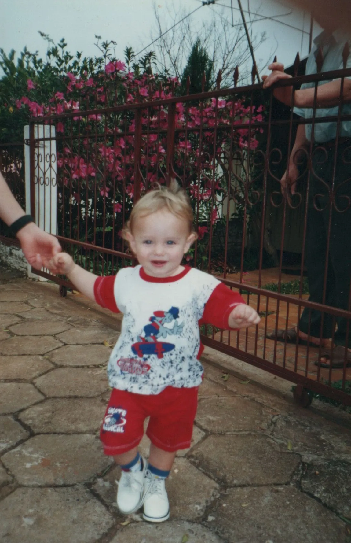 Uma criança de cabelos claros, vestindo uma camiseta estampada e shorts vermelhos, é segurada pela mão direita por um adulto enquanto caminha sorrindo em um caminho de paralelepípedos, com uma cerca de metal enferrujada coberta por flores rosa e um segundo adulto de pé ao fundo. (Original)
