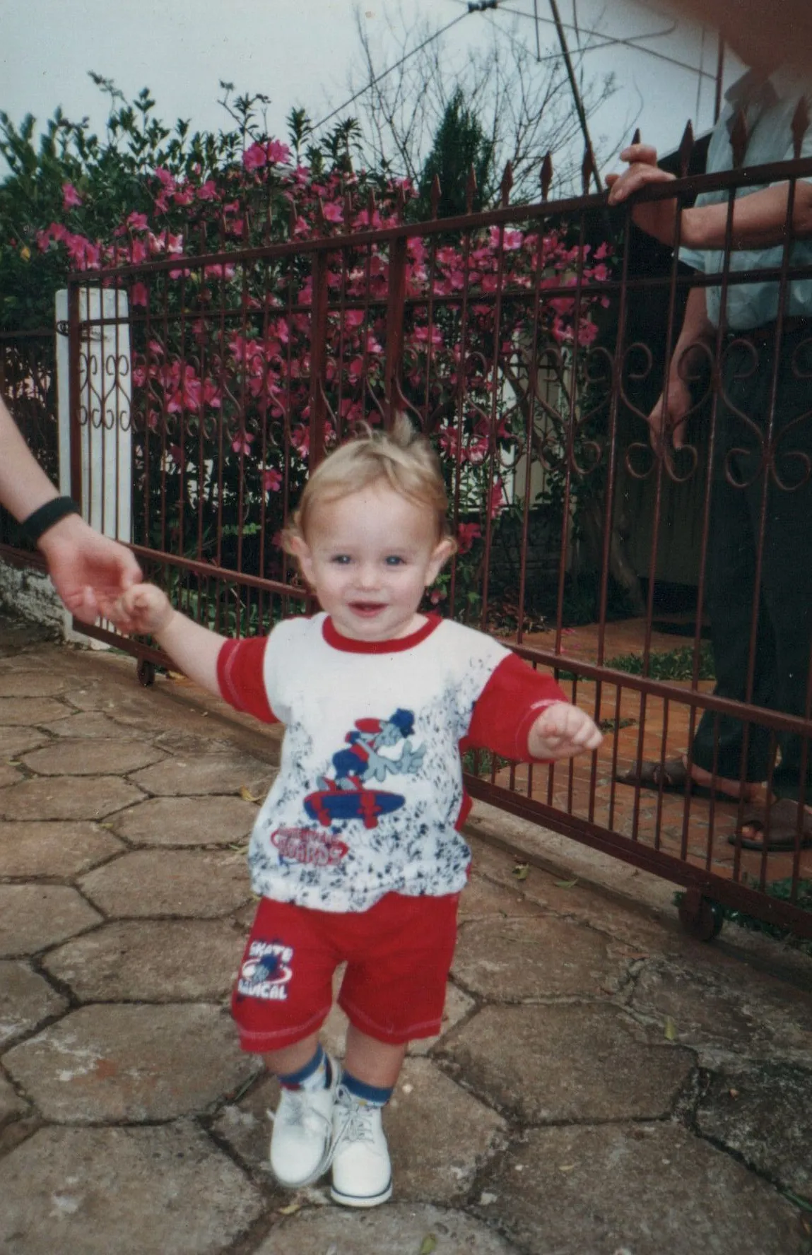 Uma criança de cabelos claros, vestindo uma camiseta estampada e shorts vermelhos, é segurada pela mão direita por um adulto enquanto caminha sorrindo em um caminho de paralelepípedos, com uma cerca de metal enferrujada coberta por flores rosa e um segundo adulto de pé ao fundo.