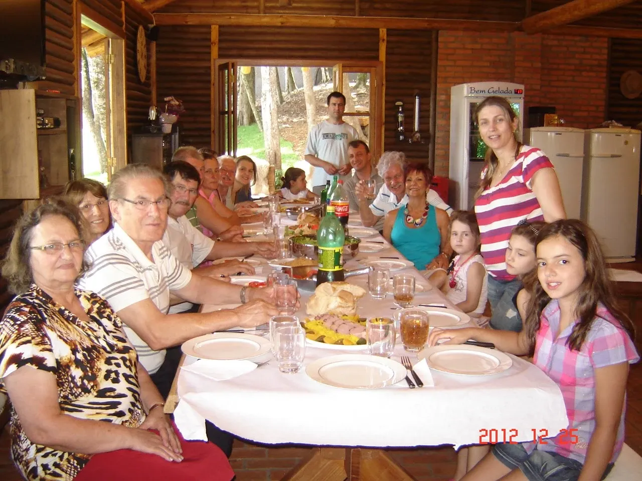 Grupo de pessoas de diferentes idades reunidas em uma longa mesa de jantar de madeira em um quarto rústico com paredes de tijolo e madeira.
