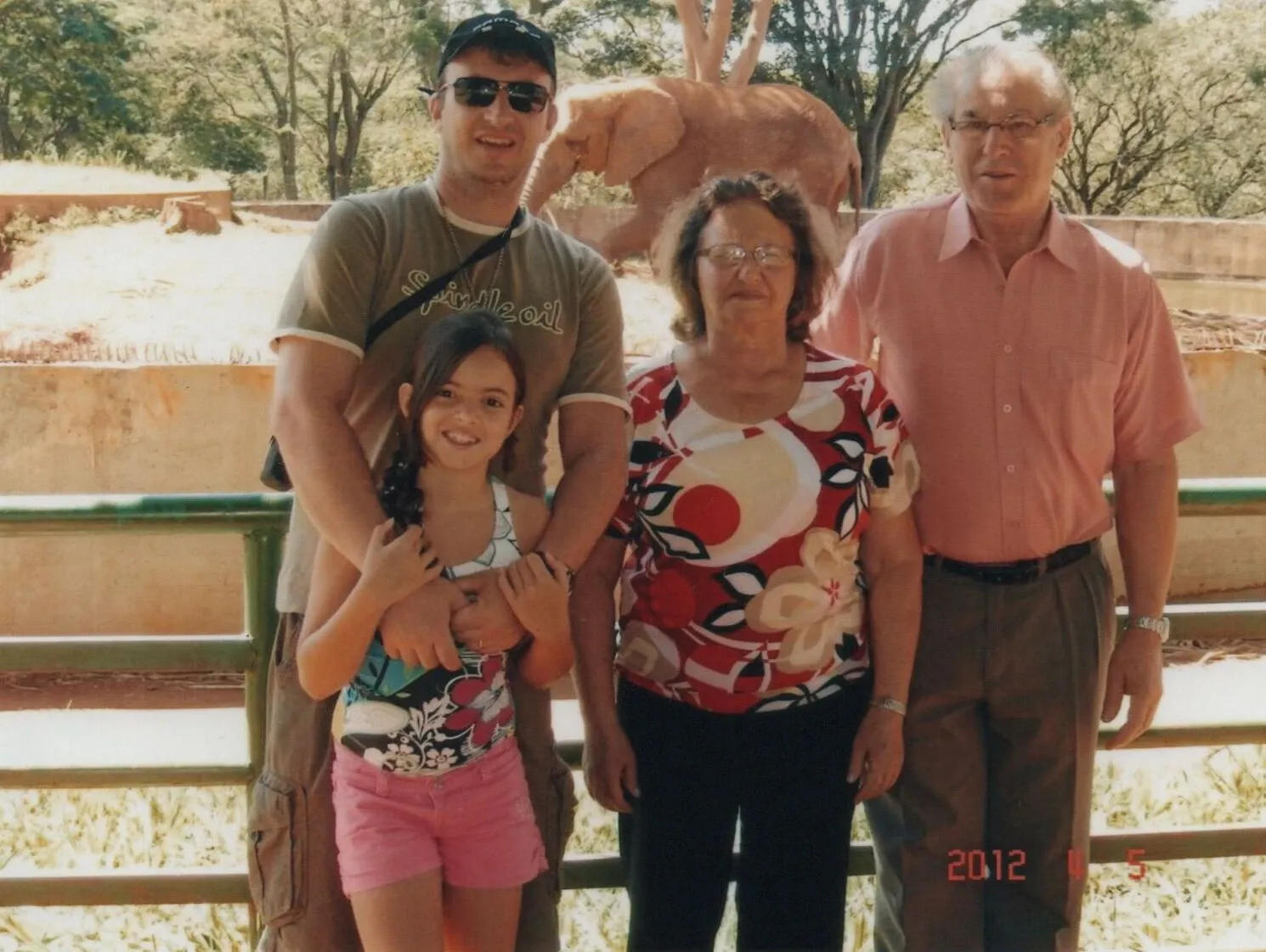 Uma família de quatro pessoas, incluindo uma menina, um homem e um casal mais velho, posa para uma foto em um zoológico. Um elefante está visível no fundo.