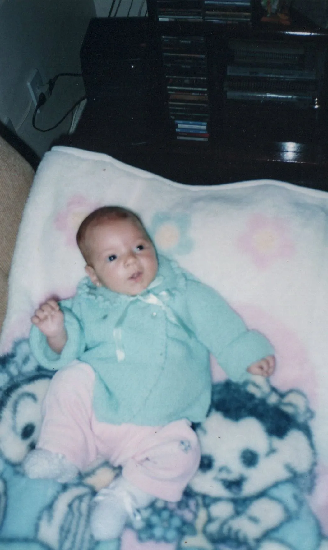 Bebê deitado em um cobertor com estampas de animais, usando um casaco de malha azul-petróleo e calças cor-de-rosa.