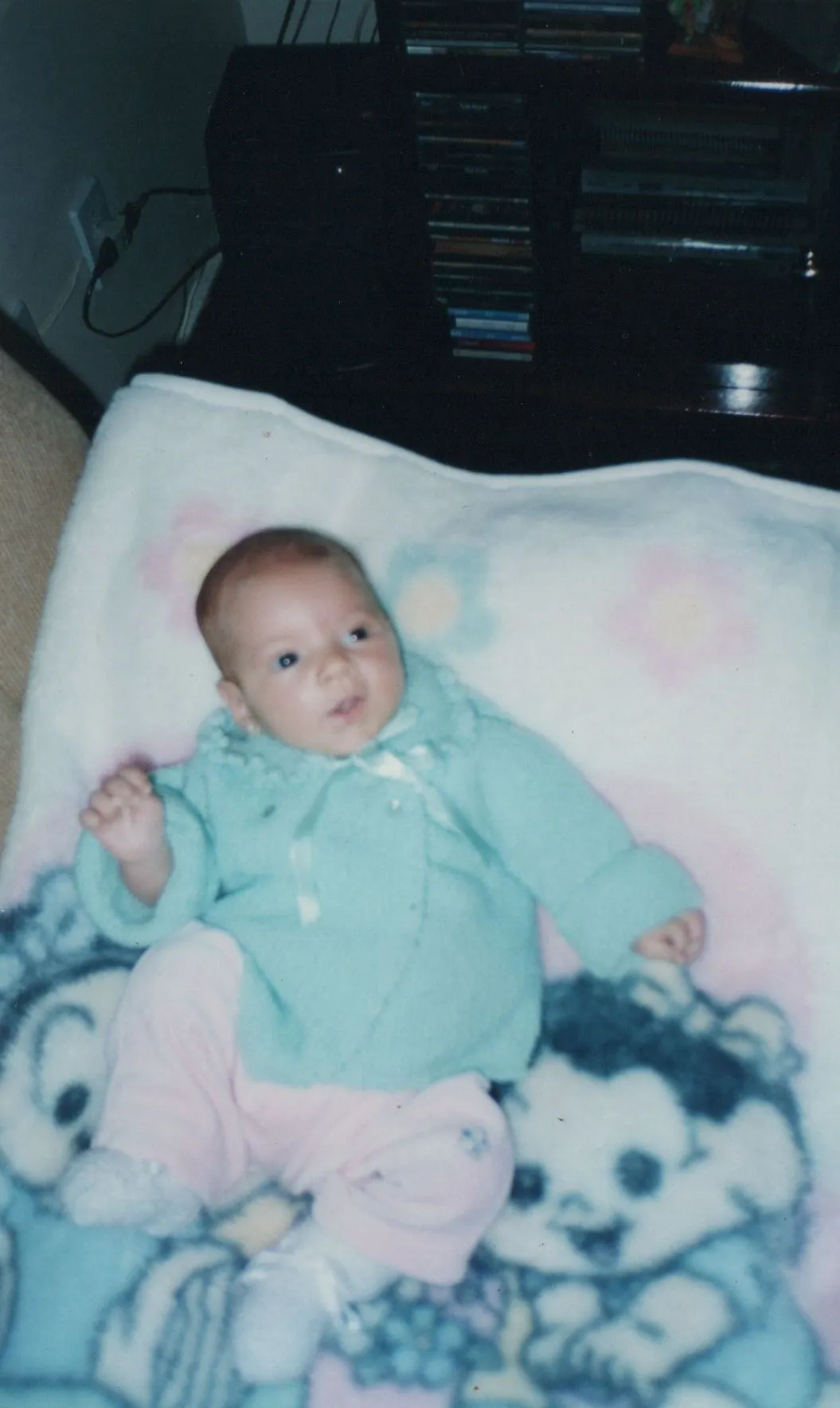 Bebê deitado em um cobertor com estampas de animais, usando um casaco de malha azul-petróleo e calças cor-de-rosa. (Original)