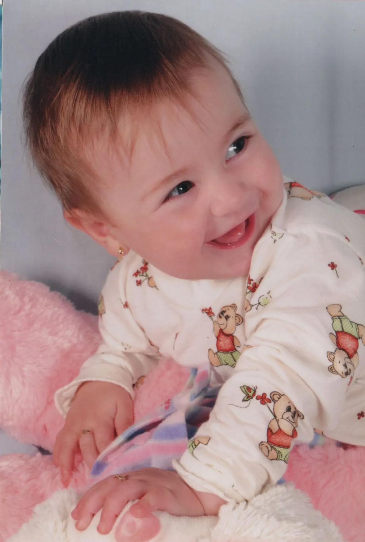Bebê de pele clara, deitado de bruços, vestindo uma camisa branca com ursinhos de pelúcia, sorrindo e olhando para a direita, com as mãos sobre um cobertor rosa e branco, e um ursinho de pelúcia branco. (Original)