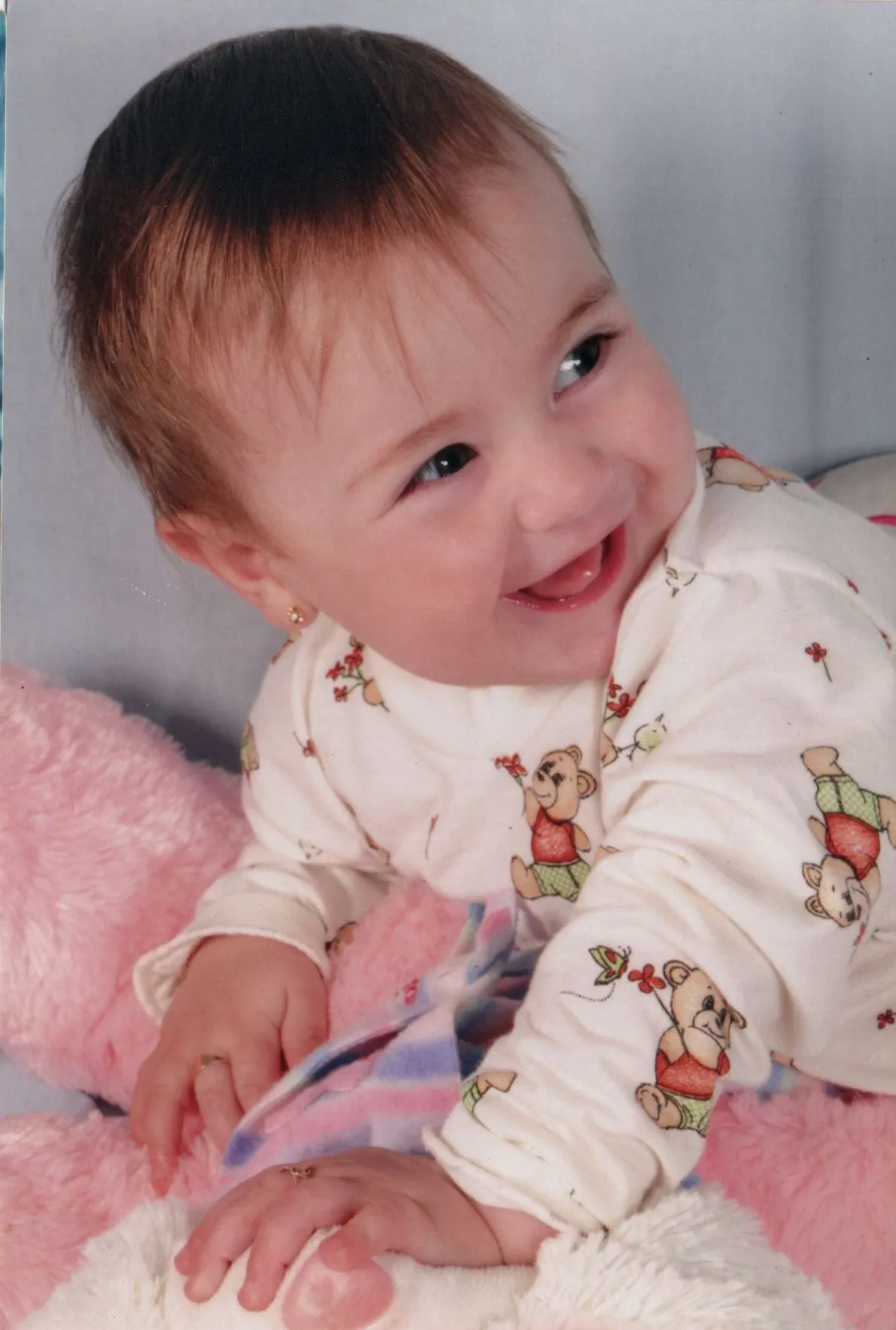 Bebê de pele clara, deitado de bruços, vestindo uma camisa branca com ursinhos de pelúcia, sorrindo e olhando para a direita, com as mãos sobre um cobertor rosa e branco, e um ursinho de pelúcia branco.